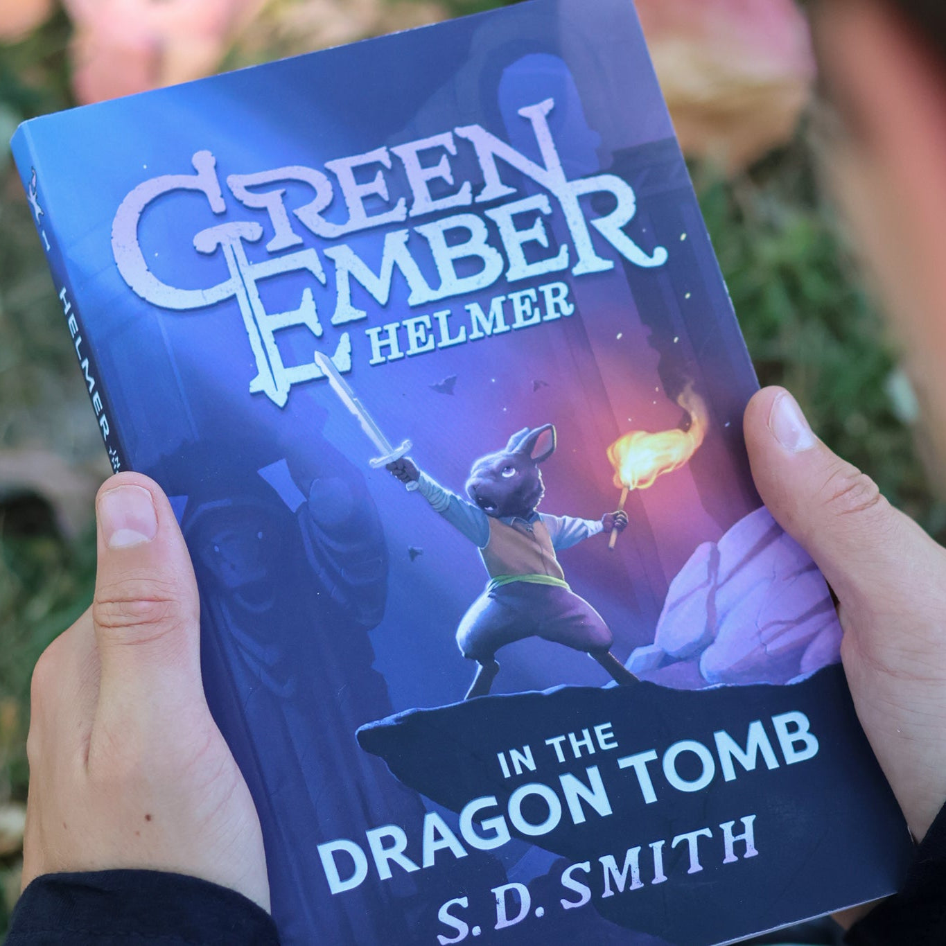 Person holding a book titled 'Green Ember Helmer in the Dragon Tomb' by S.D. Smith.