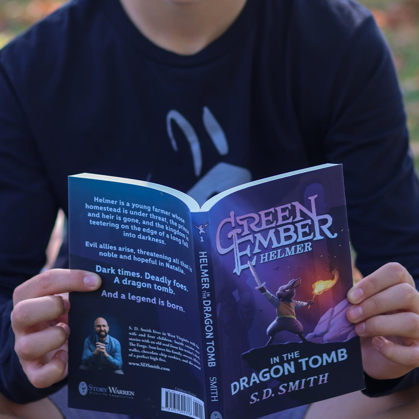 Person holding a book titled 'Green Ember Helmer: In the Dragon Tomb' outdoors.