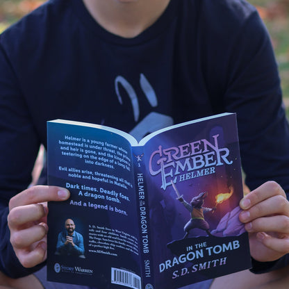 Person holding a book titled 'Green Ember Helmer: In the Dragon Tomb' outdoors.