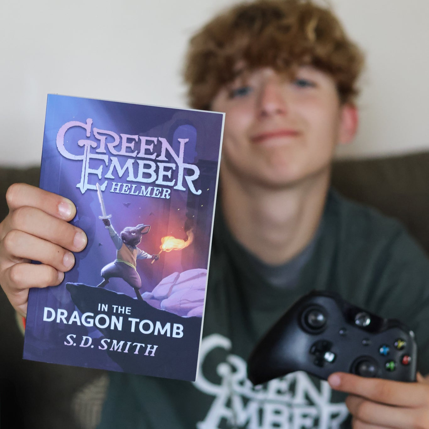 Person holding a book titled 'Green Ember: Helmer in the Dragon Tomb' by S.D. Smith, with a gaming controller.