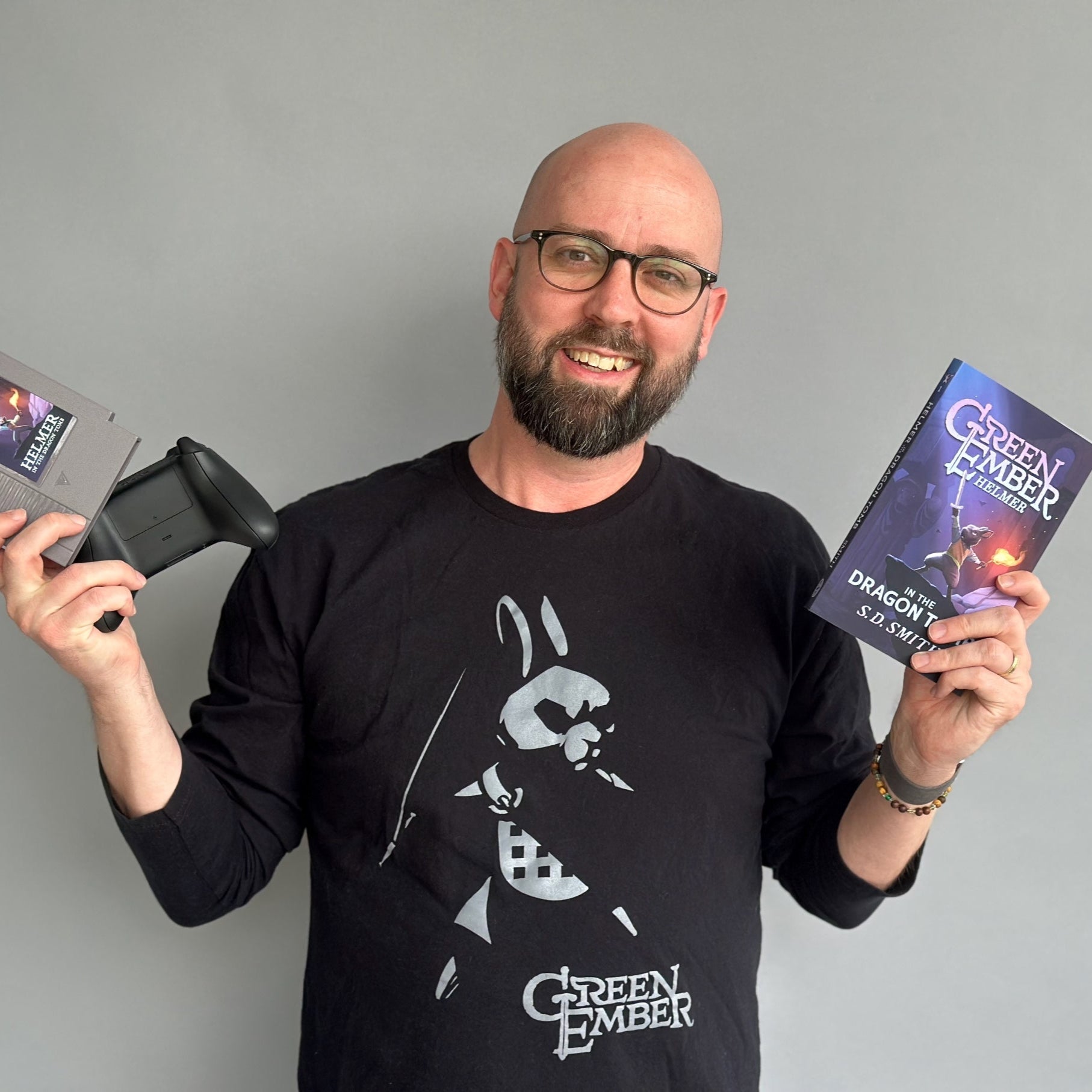 Man holding a vintage game console and a book titled 'Green Ember' against a gray background