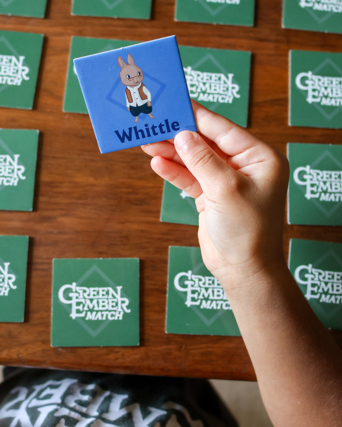 A hand holds a custom blue square coaster featuring a cartoon rabbit and the name 'Whittle' in white text. The background shows a wooden table covered with green square coasters printed with the words 'GREEN EMBER MATCH'.